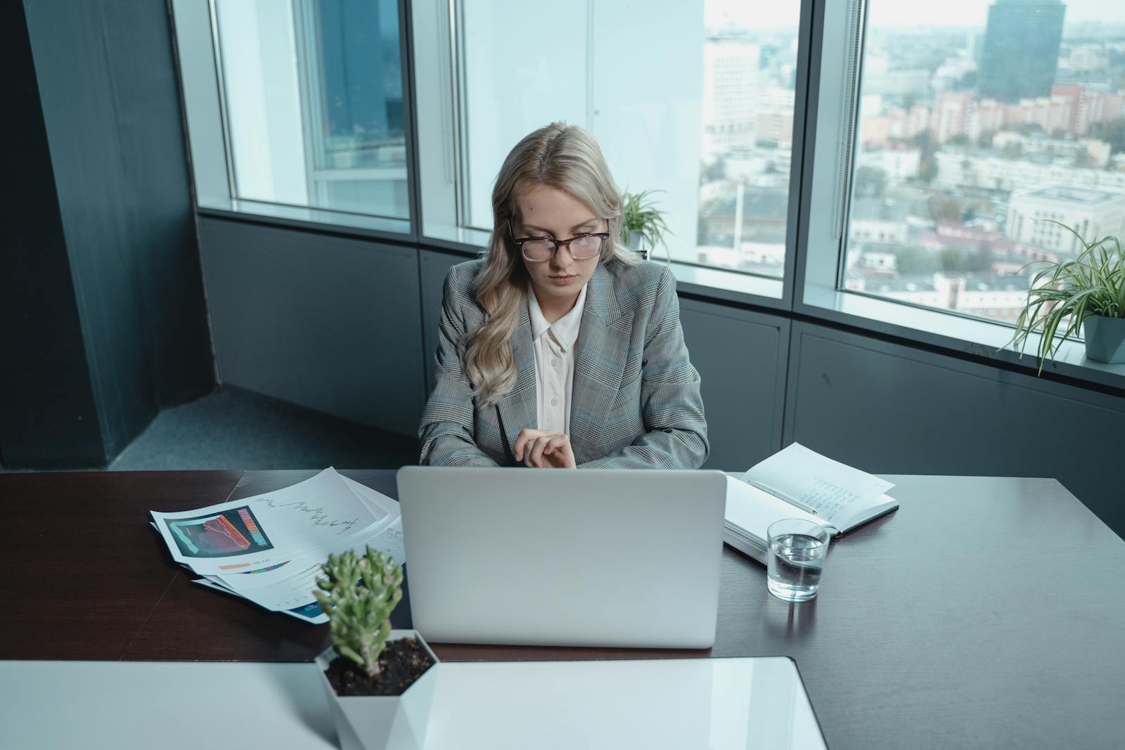 Professional woman in office setting focused on work with MacBook and documents.
