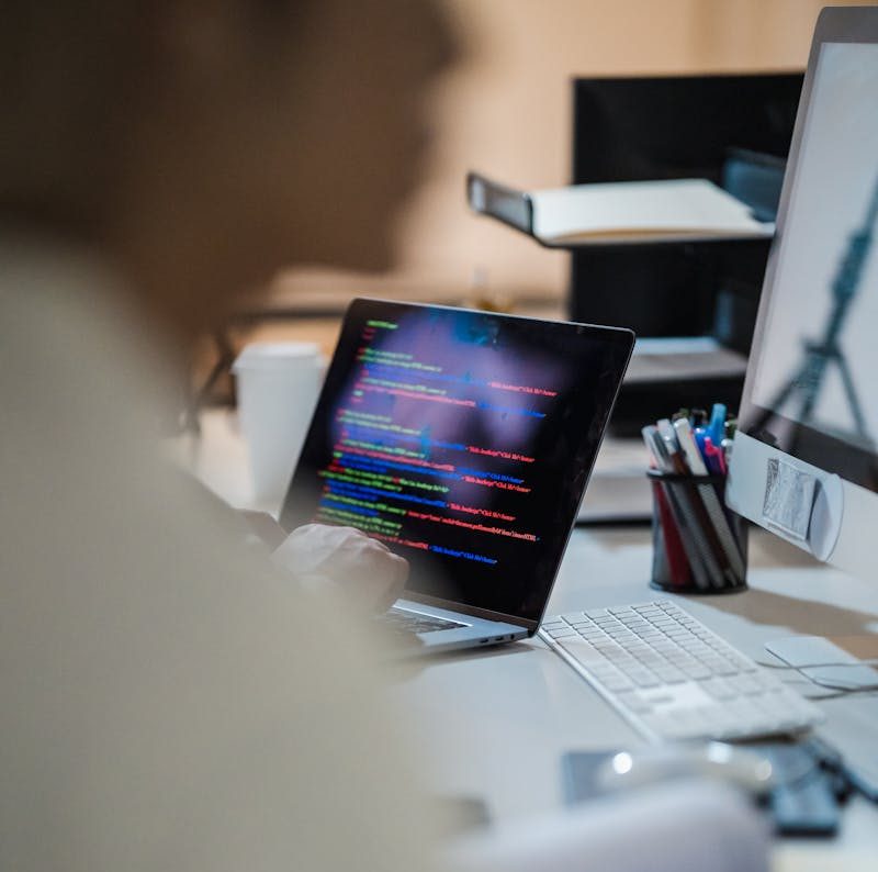 Blurred figure at a desk with a laptop displaying colorful code, indicating a programming environment.